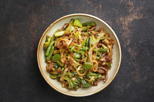 Fideos orientales, vista superior. Carne frita en un wok con fideos de arroz, judías verdes, calabacín, pak choi y pimientos verdes en salsa picante de soja-ostra, en un tazón, sobre fondo pedregoso negro. Plato de cousine tailandés. — Foto de Stock