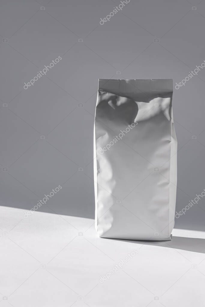 Blank white foil pouch bag on grey background in minimal style with natural hard shadows. Monochrome food photo. Packaging valve and seal template mockup. Metallic coffee tea retail package design.