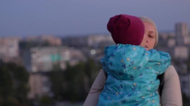 Anne ve kızı şehir manzarasında duruyor. Gün batımında annesini kucaklayan ve öpen küçük bir kız. Sevgi dolu çocuk annesine sarılıyor. Anneler Günü aile karşılama kartı. Yaşam tarzı 4K