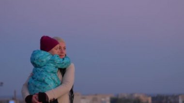 Anne ve kızı şehir manzarasında duruyor. Gün batımında annesini kucaklayan ve öpen küçük bir kız. Sevgi dolu çocuk annesine sarılıyor. Anneler Günü aile karşılama kartı. Yaşam tarzı 4K