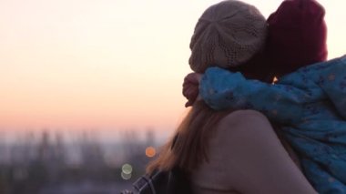 Anne ve kızı şehir manzarasında duruyor. Gün batımında annesini kucaklayan ve öpen küçük bir kız. Sevgi dolu çocuk annesine sarılıyor. Anneler Günü aile karşılama kartı. Yaşam tarzı 4K