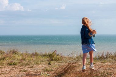 Kot ceketli sevimli küçük kız deniz manzaralı bir tepenin arkasını görüyor. Kırsal alanda bulutlu arka planda uzun sarı saçlı şık bir çocuk. Yüzü olmayan kırsalda gezintiye çıkmak..