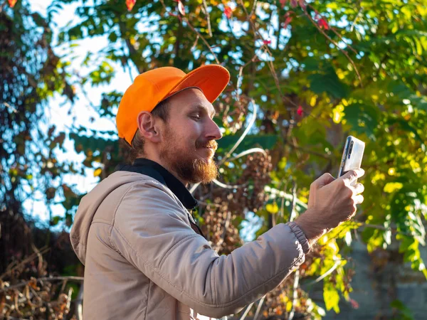 Young red bearded man speaks on mobile phone smiles. Guy in raincoat and orange cap on colorful autumn background. Positive emotional businessman answers smartphone call. Lifestyle work communication.