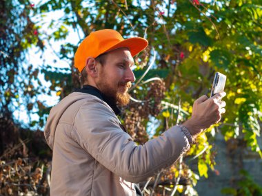 Genç kızıl sakallı adam cep telefonuyla gülümsüyor. Renkli sonbahar arka planında turuncu şapkalı bir adam. Pozitif duygusal iş adamı akıllı telefon çağrılarına cevap verir. Yaşam tarzı iş iletişimi.