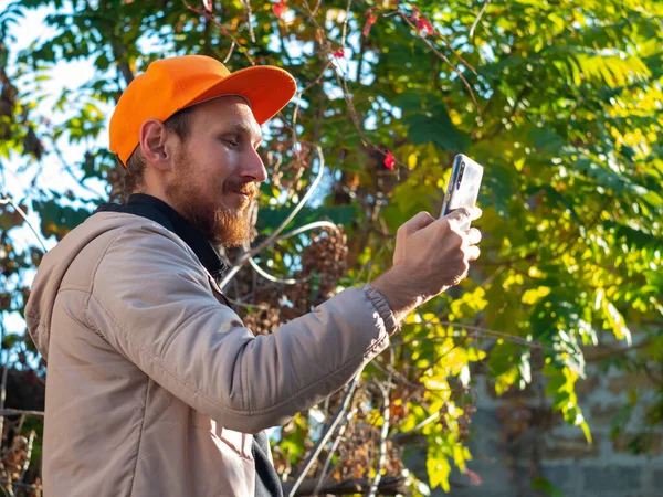 Genç kızıl sakallı adam cep telefonuyla gülümsüyor. Renkli sonbahar arka planında turuncu şapkalı bir adam. Pozitif duygusal iş adamı akıllı telefon çağrılarına cevap verir. Yaşam tarzı iş iletişimi.