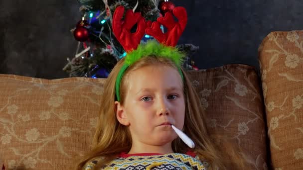 Szomorú, beteg gyerek lázmérővel a szájában szilveszterkor. Aranyos kislány ünnepi vörös szarvas szarvakkal sír könnyes szemekkel. Depressziós vírus betegség beteg ideges boldogtalan gyerek ünnepli a karácsonyt