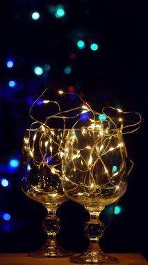 Noel masasında altın renkli çelenk bulunan kristal şarap kadehleri arka plandaki dikey videoyu aydınlatıyor. Romantik yeni yıl arifesi aşıklar için şampanyası. Sihirli kış tatili atmosferi