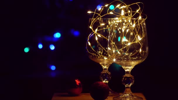 クリスマステーブルのボケの照明の背景に金色の輝くガーランドで飾られたクリスタルワイングラス ロマンチックな新年の愛のカップルのためのシャンパンと夕食を前夜 魔法の冬の休日の雰囲気 — ストック動画