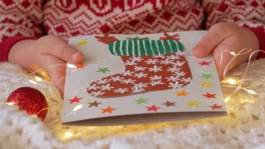 Noel çorabı el yapımı tebrik kartı projesi. Kırmızı Noel Baba hediye çantalı renkli kart. Çocuk, beyaz örülü ekose arka planda ışıklandırmalı Noel şenliği süslemeleri yapıyor..