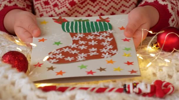 クリスマスソックス手作りグリーティングカードDiyプロジェクト 赤いサンタストッキングキャンディーギフトバッグとカラフルなカード 子供は新年のお祝いの装飾を作る白いニットの背景にライトで遊ぶ — ストック動画