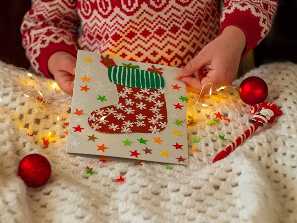 Christmas sock handmade greeting card DIY project. Colorful card with red Santa stocking candy gift bag. Kid makes New Year holidays festive decorations on white knitted plaid background with lights.