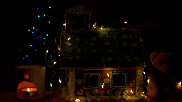 柔らかいクマと自家製の甘いジンジャーブレッドハウス アロマランプで輝くキャンドルディフューザーお祝いのガーランドの装飾がぼやけてクリスマスツリーボケライト暗い背景 — ストック動画