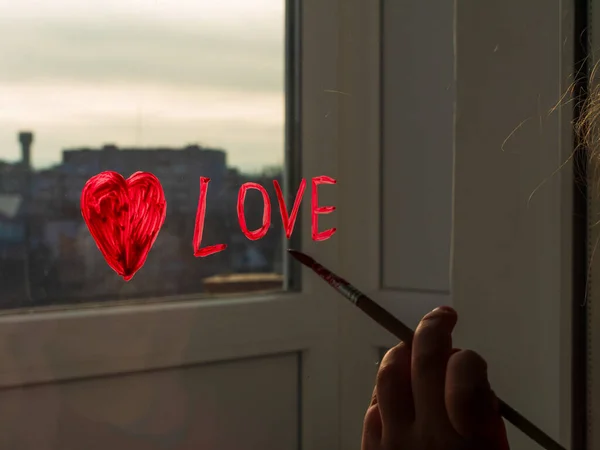 Little girl holds paintbrush in hand and drawing red heart on window glass. Valentine's day, love, dating, quarantine family leisure. Stay home art concept. Child painting playing indoors. New normal.