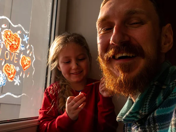 Child with father painting Love sign, playing indoors quarantine family leisure. Little girl holds paintbrush in hand draws red heart on window glass. Valentine\'s day. Stay home art concept New normal