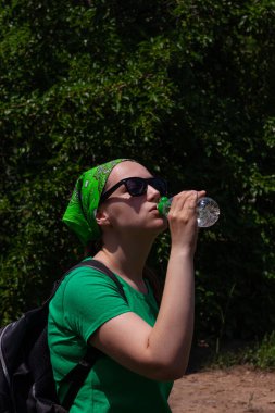 Genç kız elinde bir şişe su tutuyor ve içiyor. Güneş gözlüklü, yeşil gömlekli, bandanalı güzel hippi portresi güneşli bir günde doğada duruyor. Kilo kaybı, yaz mevsiminde aktif vücut bakımı.