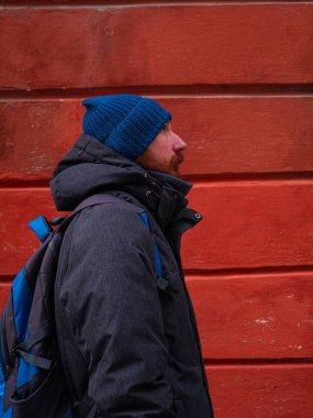 Mavi şapkalı, gri ceketli, sırt çantalı, kırmızı duvarlı sakallı adam. Şehir tarzı sokak gezisi fotoğrafı. Dışarıda duran bıyıklı mutlu, yakışıklı hippi adam. Etkin yaşam tarzı yürüyüşü kavramı.