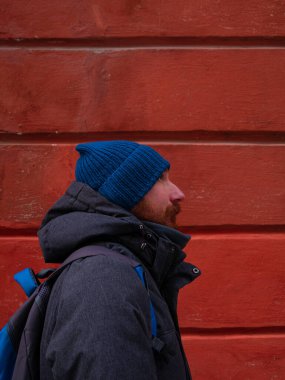 Mavi şapkalı, gri ceketli, sırt çantalı, kırmızı duvarlı sakallı adam. Şehir tarzı sokak gezisi fotoğrafı. Dışarıda duran bıyıklı mutlu, yakışıklı hippi adam. Etkin yaşam tarzı yürüyüşü kavramı.