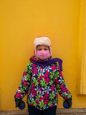 Sarı duvar arkasında koruyucu pembe maske takan kalın kışlık elbiseli genç bir kız. Şehir tarzı sokak fotoğrafı. Dışarıda tek başına duran mutlu çocuk okulu. Yeni normal güvenlik yaşam tarzı konsepti..