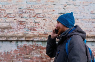 Genç kızıl sakallı adam cep telefonuyla konuşuyor mavi şapkalı gri ceketli adam tuğla duvarlı arka planda olumlu duygusal bin yıllık cevaplar akıllı telefon çağrısı. Yaşam tarzı iş mesafesi iletişimi.