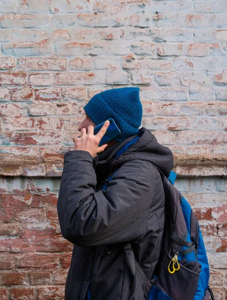 Young red bearded man speaks on mobile phone smiles Guy in blue hat gray coat on brick wall urban background Positive emotional millennial answers smartphone call.Lifestyle distance work communication