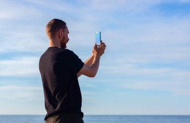 Denizde siyah tişörtlü sakallı bir adam ve açık mavi gökyüzü manzarası arka planı elinde akıllı telefonuyla fotoğraf çekiyor. Genç adam cep telefonuyla doğayı fotoğraflıyor, aktif yaşam tarzı seyahat kavramı.