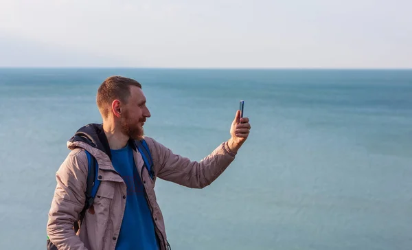 Bearded man in raincoat with backpack on sea landscape with blue cloudy sky background with smartphone in hand taking picture. Guy photographing nature on cell phone, active lifestyle travel concept.