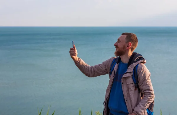 Bearded man in raincoat with backpack on sea landscape with blue cloudy sky background with smartphone in hand taking picture. Guy photographing nature on cell phone, active lifestyle travel concept.
