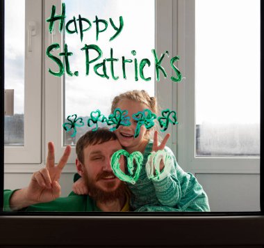 Üç yapraklı yeşil yoncalar, şenlikli ev dekorasyonu ve aile eğlencesi çizen kızıyla Aziz Patrick Günü Baba 'yı çizmek. Pencere camına yonca yaprağı çizmek. Ev konseptini koru Yeni normal