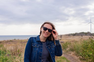 Tepede kot ceketli, yabani çimenler ve mavi deniz altyapısı olan bir kadın. Kırsal alanda, bulutlu gökyüzü okyanus manzarasında uzun esmer saçlı şık bir kız. Kendine güveni tam bir kadın kırsal alanda geziniyor..