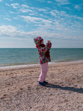 Boş deniz sahilinde fotoğraf çeken bir çocuk. Kalın elbiseli küçük kız elinde kamerayla fotoğraf çekiyor. Yaşam tarzı fotoğraf Soğuk hava bahar mevsimi Aile eğlencesi tatil tatili reklamı