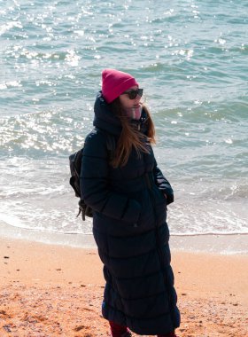 Pembe şapkalı, siyah güneş gözlüklü ve sıcak elbiseli genç bir kadın kumsalda mavi deniz suyuyla yürüyor. Dış mekan tarzı otantik bir fotoğraf. Tek başına seyahat eden gerçek insanlar.