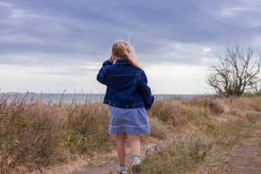 Kot ceketli sevimli küçük kız deniz manzaralı bir tepenin arkasını görüyor. Kırsal alanda bulutlu arka planda uzun sarı saçlı şık bir çocuk. Yüzü olmayan kırsalda gezintiye çıkmak..