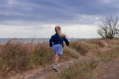 Kot ceketli sevimli küçük kız deniz manzaralı bir tepenin arkasını görüyor. Kırsal alanda bulutlu arka planda uzun sarı saçlı şık bir çocuk. Yüzü olmayan kırsalda gezintiye çıkmak..