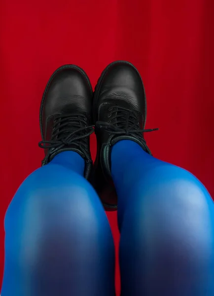 Female legs in blue tights and black boots upside down on red background. Spring autumn fashion collection modern style. Vivid vibrant contrasting colors. Fashionable advertising vertical banner.