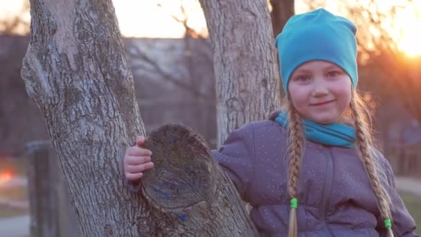 在春天的公园里 一个快乐的女孩 留着长长的辫子 坐在一棵落日下的树上 秋天的森林里 穿着时髦外套头戴帽子的孩子在夕阳西下的时候笑着拍照 — 图库视频影像