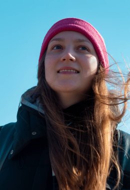Pembe şapkalı, uzun kahverengi saçlı, kendine güvenen genç bir kadın açık mavi gökyüzü arkaplanı. Milenyum kızı, güneşli bir günde dışarıda geziyor. Aktif yaşam tarzı, otantik fotoğraf. Tek başına seyahat eden gerçek insanlar.