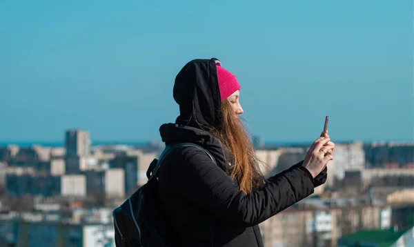 年轻自信的女人戴着粉红的帽子 手里拿着智能手机 给在城市背景蓝天上散步的朋友打电话 时尚的千禧年女孩户外生活方式正宗照片 真正的人 独自旅行 — 图库照片