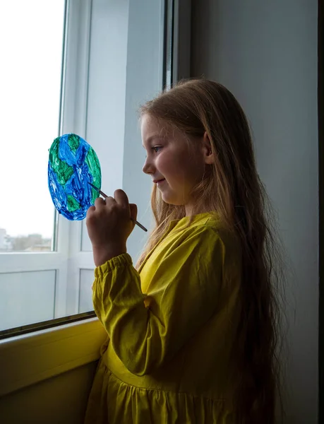 Cute little girl painting planet on window at home. Happy Earth Day April 22 greeting message. Creative family leisure lockdown new reality. Ecology Saving environment conscious consumption concept.