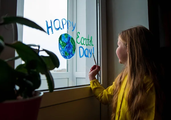 Cute little girl painting planet on window at home. Happy Earth Day April 22 greeting message. Creative family leisure lockdown new reality. Ecology Saving environment conscious consumption concept.