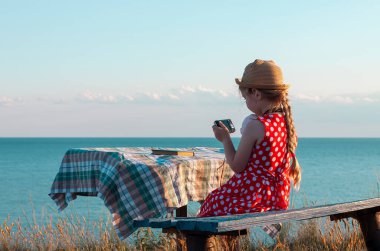Hasır şapkalı küçük fotoğrafçı çocuk ve klasik bankta kırmızı benekli elbise deniz manzarasında yumuşak pembe tavşan oyuncağının fotoğrafını çekiyor. Kız elindeki kameraya bakıyor. Arkadaşlık eğlence seyahati.