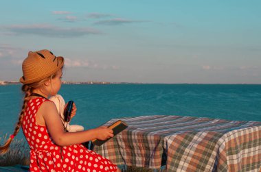 Hasır şapkalı küçük fotoğrafçı çocuk ve klasik bankta kırmızı benekli elbise deniz manzarasında yumuşak pembe tavşan oyuncağının fotoğrafını çekiyor. Kız elindeki kameraya bakıyor. Arkadaşlık eğlence seyahati.