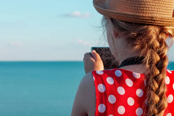 年轻的摄影师小孩戴着草帽 穿着红色圆圆点裙 坐在老式长椅上 拍摄着海景背景下柔软粉色兔子玩具的照片 女孩手里拿着相机 友谊旅途愉快 — 图库照片