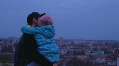 Baba ve kızı akşam kentinde yürürken gülümsüyor. Yaşam tarzı gerçek insanlar. Mor günbatımı gökyüzü olan şehir manzarası. Mutlu aktif aile alacakaranlıkta eğleniyor, yeni normal sosyal uzaklık.