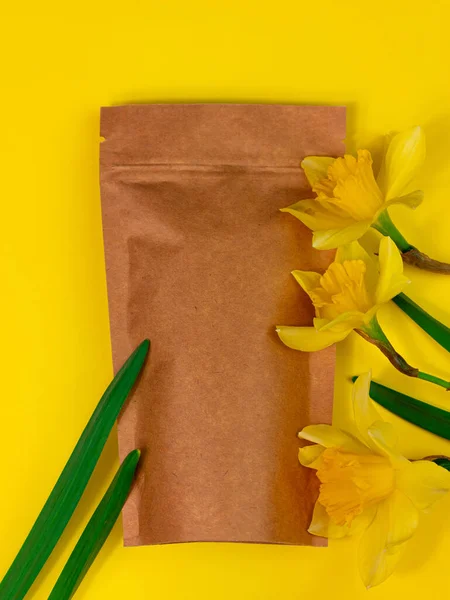 Brown kraft paper pouch bag mockup coffee beans and yellow daffodil flowers on pastel background top view. Packaging for food product template.Pack with clasp for tea leaves,weight flat lay copy space