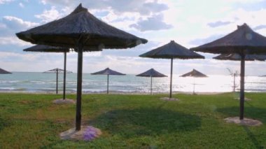 Yeşil çimenler üzerinde saman şemsiyeler, kumsal, canlı mavi deniz suyu ve güneş ışınlarında gökyüzü arkaplanı. Sahne manzarası 4K görüntüler, yaz tatili mevsimlik tropikal deniz seyahati dinlenme, tatil beldesi reklamı..