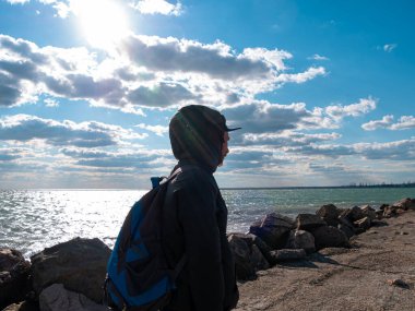 Mavi bulutlu arka planda, gür sakallı ve bıyıklı yakışıklı hippi. Sırt çantalı, siyah yağmurluk giyen kızıl sakallı adam. Ciddi vahşi erkek yalnız. Boş deniz kıyısında şık bir gezgin