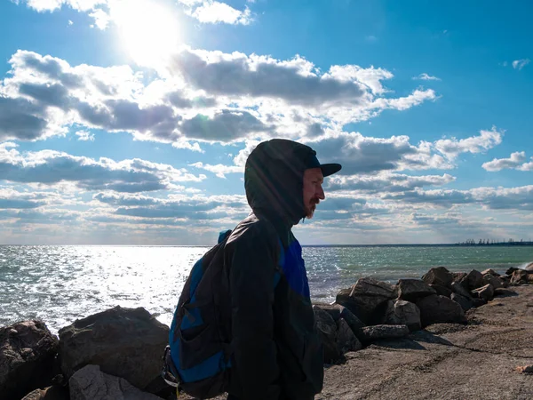 Handsome hipster man with lush beard and mustache on blue cloudy sky background. Red bearded guy in black raincoat with backpack outdoor. Serious brutal male alone. Stylish traveler on empty sea shore