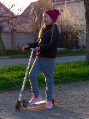 Çocuk tek başına kickscooter kullanıyor. Spring City Park sokağında motosikletli sarışın tatlı kız. Aktif yaşam tarzı, spor eğitimi. Yeni normal etkinlik kilitleme sosyal mesafesi.