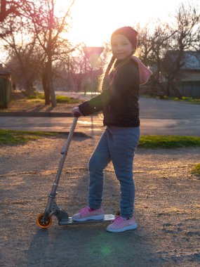 Çocuk tek başına kickscooter kullanıyor. Spring City Park sokağında motosikletli sarışın tatlı kız. Aktif yaşam tarzı, spor eğitimi. Yeni normal etkinlik kilitleme sosyal mesafesi.