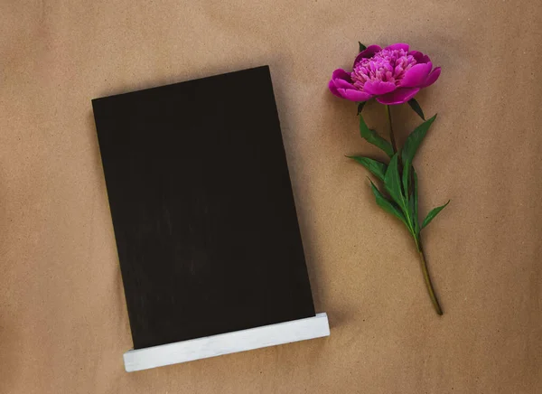 Blank chalkboard mockup with pink peony on brown kraft paper background. Blackboard with beautiful flower wallpaper copy space. Minimalism style. Birthday greeting card floral gift template text sign.